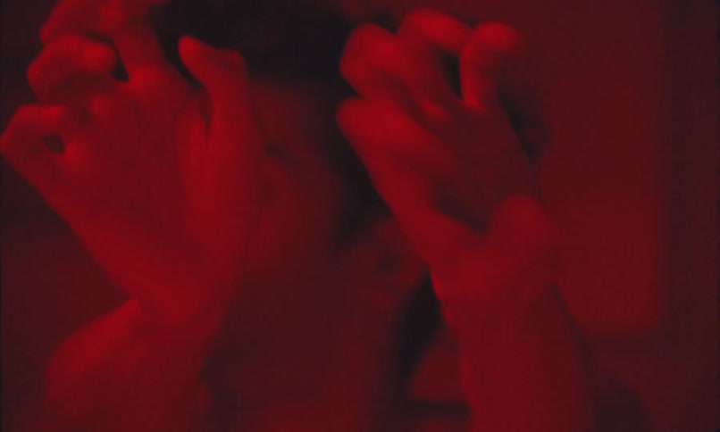 Man lying on his back in a red lit room awkwardly twists his wrists repeatedly