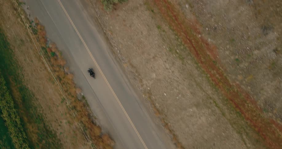Aerial shot of a lone motorcycle riding down a road cutting between farmland and dry terrain