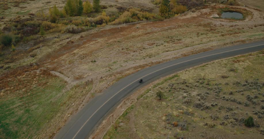 A lone motorcyclist rides along a winding rural road captured from an aerial view