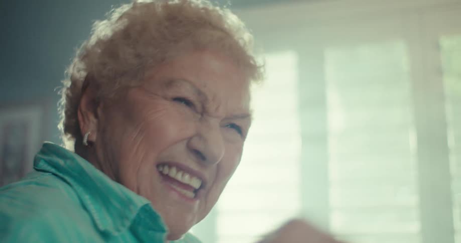 Elderly woman talking as she dances in front of a sunlit window in her living room