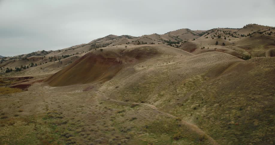 An expansive aerial view showcases undulating earthy hills with sparse trees and patches of dry vegetation under an overcast sky