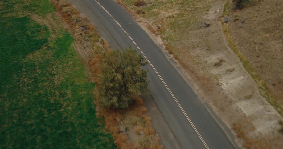 An aerial view captures a lone motorcyclist riding along an asphalt road that winds through brown hills and green fields beneath an overcast sky