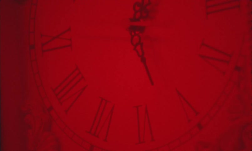 A square analog wall clock hangs on a wall in a room illuminated by red light