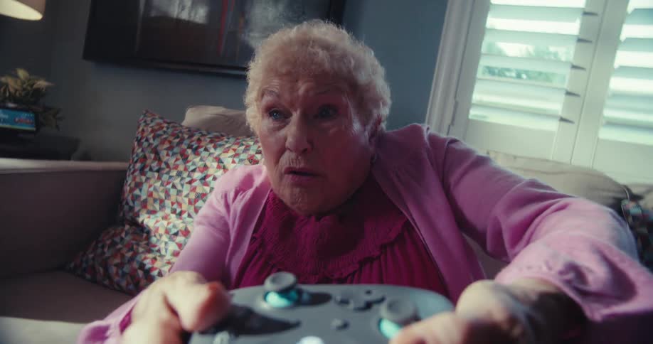 An elderly woman anxiously tilts from side to side while gaming with a controller in a living room