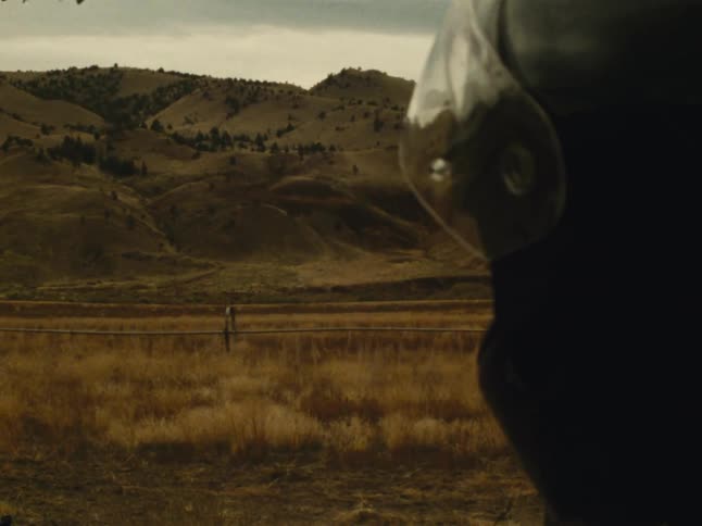 A person wearing a black helmet looking at a hill with a large beige grass field in front