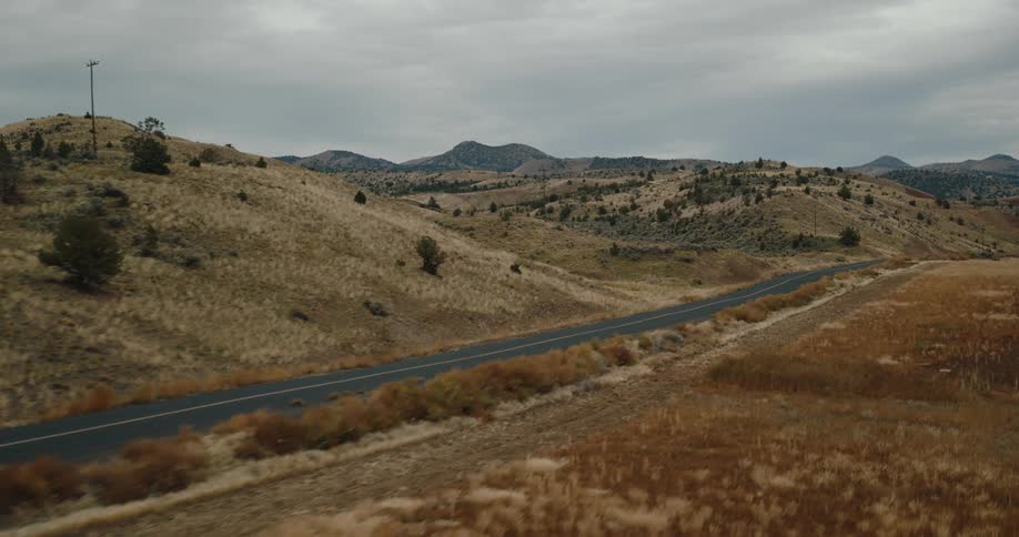 An aerial shot flies alongside a paved road that winds through a vast rural landscape of dry rolling hills under a cloudy sky