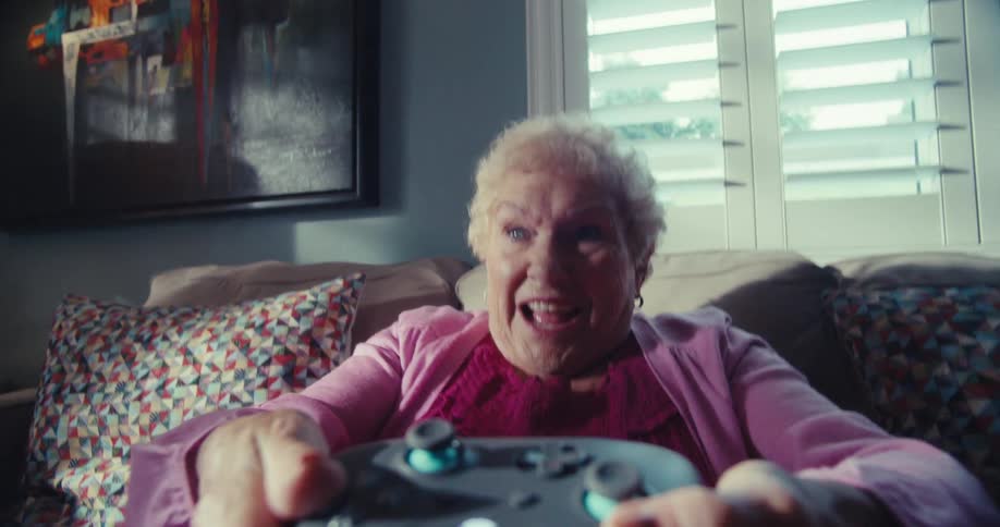 An elderly woman with short blonde hair sits on a couch her face lit with excitement as she enthusiastically plays a video game with a controller