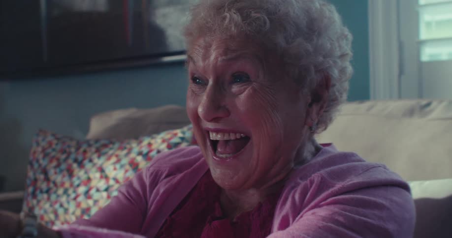 An elderly woman laughs excitedly while gaming with a controller on a couch indoors