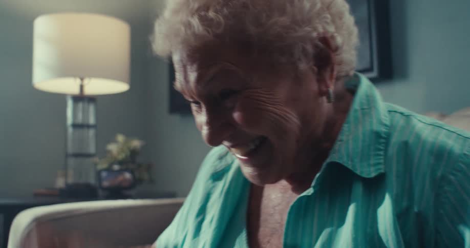 The face of an elderly woman wearing a blue shirt who is sitting on a sofa and is very happy celebrating