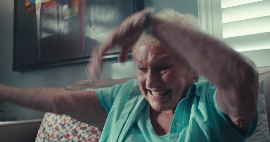 An elderly woman on a couch cheers joyfully while looking at a tablet