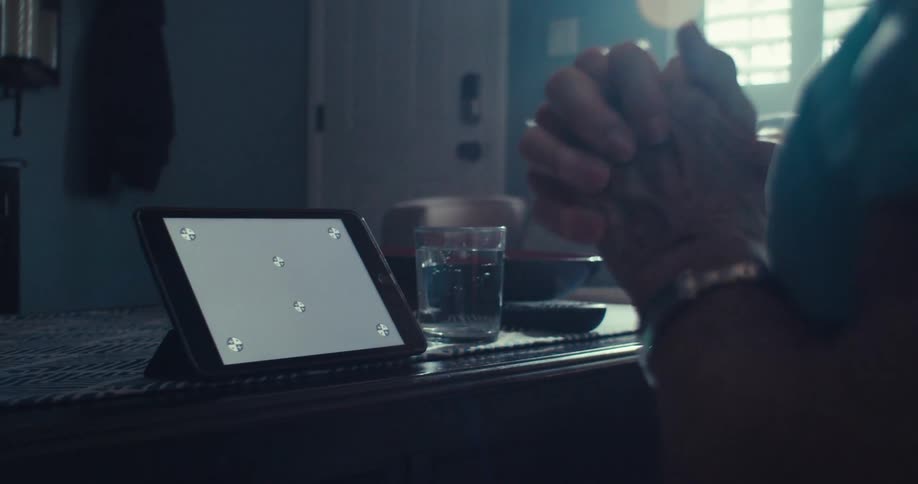 An elderly woman in a blue shirt is sitting looking at the screen of a tablet and she is crossing her hands nervously