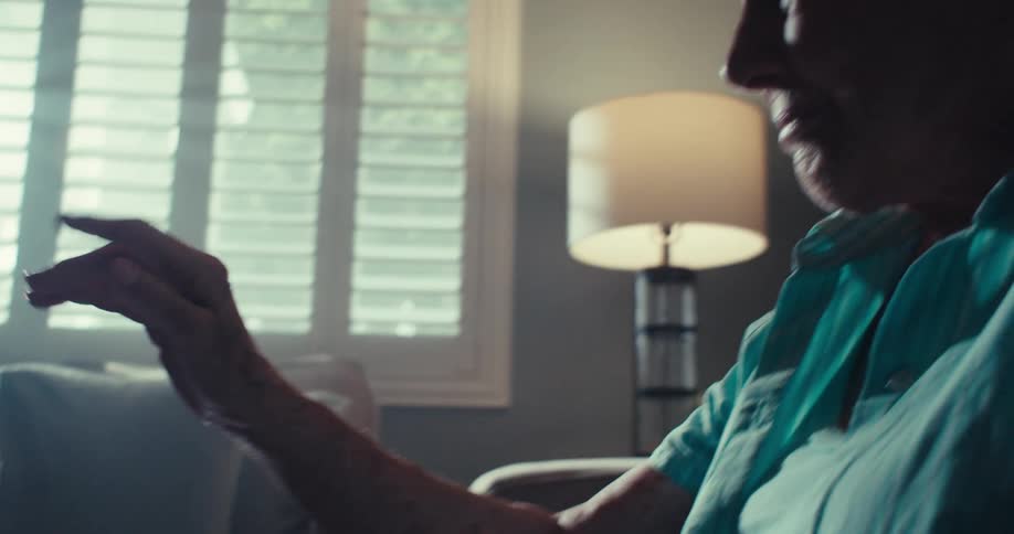 An elderly woman is on a video call using a tablet pc placed on a table smiling and waving her hand over it