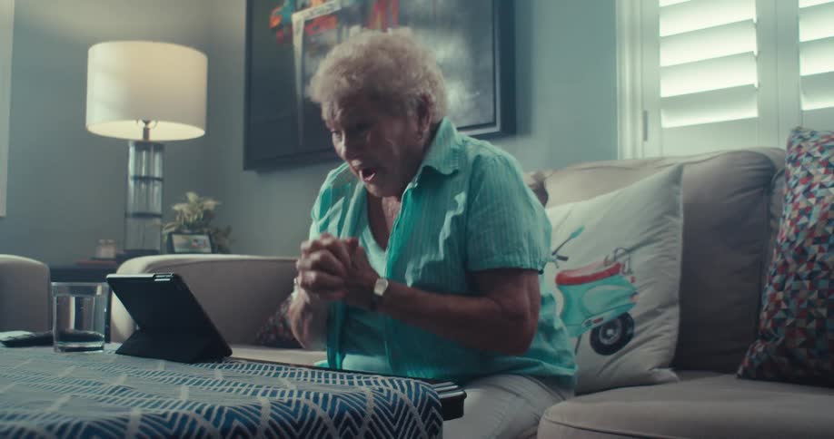 An elderly woman is joyfully laughing while using a tablet in a cozy living room