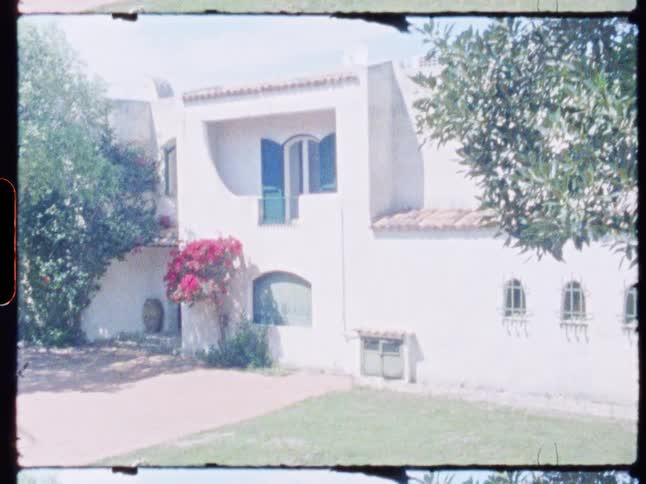 White mediterraneanstyle house with green shutters under bright sun partly shaded by trees and flowers