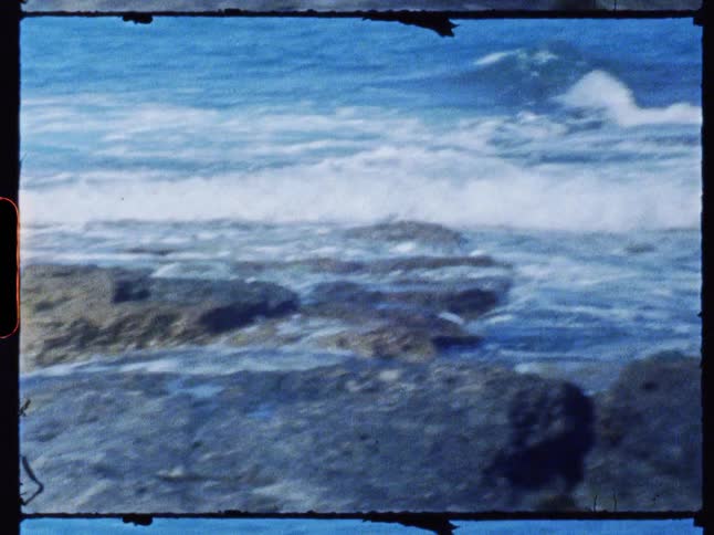 Archival footage with a visible film border shows ocean waves washing over a rocky shoreline on a sunny day