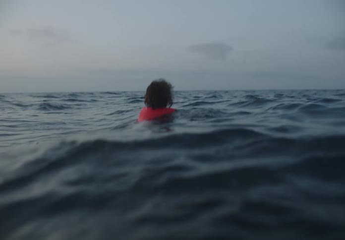 A lone child seen from behind floats in choppy dark blue water wearing an orange life vest under a cloudy overcast sky