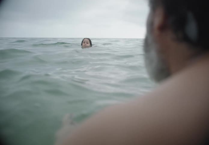 Older man floats while a child swims toward him on an overcast day
