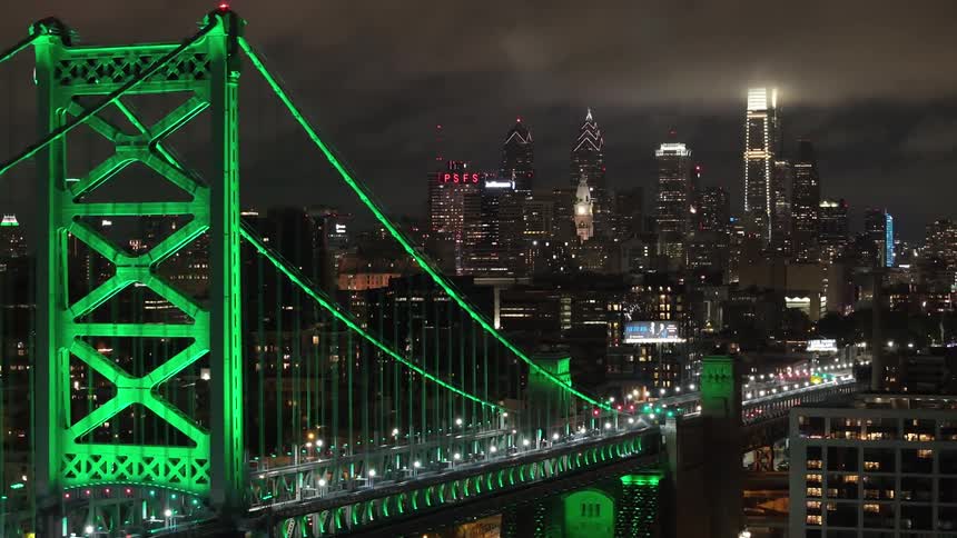 An aerial shot moves toward a downtown Philadelphia skyline at night passing by Benjamin Franklin Bridge illuminated with vibrant green lights