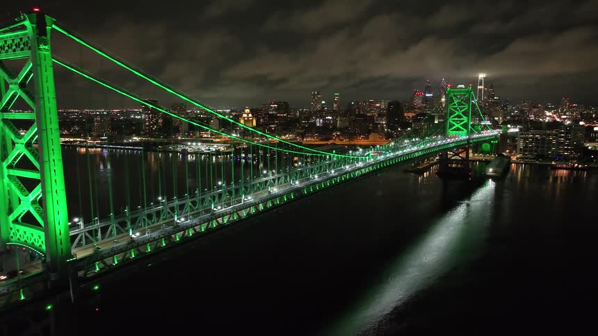 A stunning aerial view captures Benjamin Franklin Bridge in Philadelphia illuminated in vibrant green as it spans a river at night with a glowing city skyline in the background