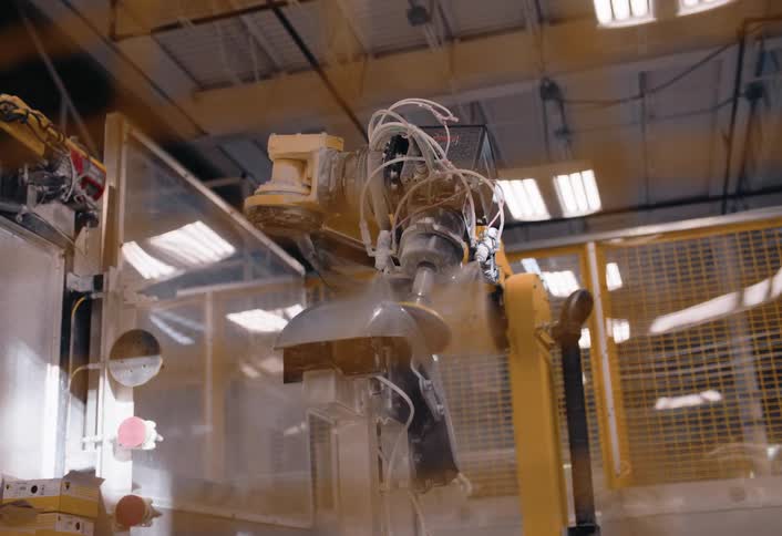 A robotic arm sprays paint onto a small object mounted on a platform in an industrial workspace