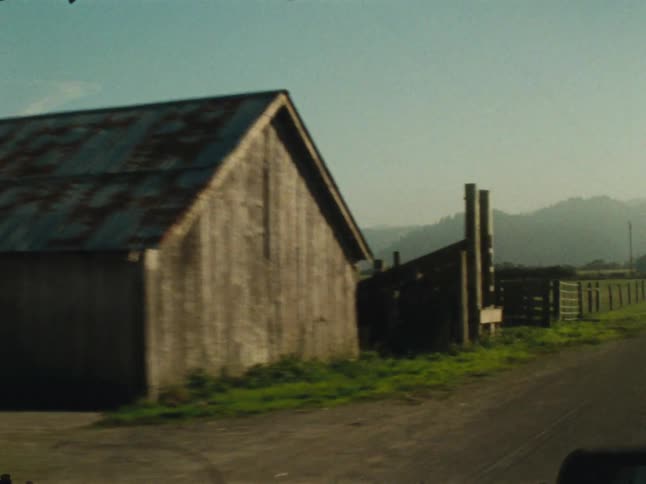 Old buildings and an open green pasture pass down a country road on a cloudy day
