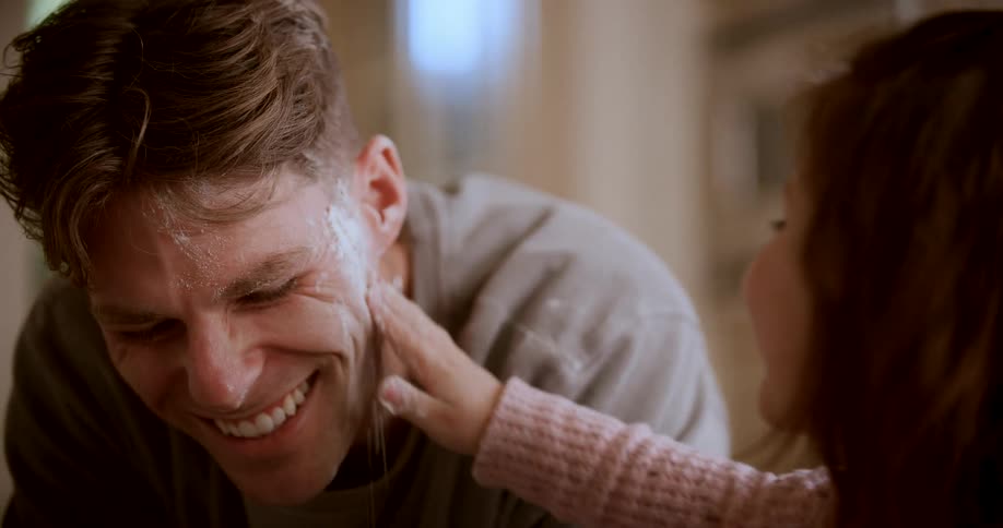 A kid rubs flour on her fathers face as he smiles and laughs both enjoying time in the kitchen at midday