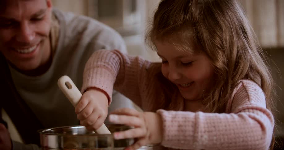 A kid stirs food in a pot while laughing as her dad stands beside her also laughing and encouraging her both enjoying a fun time in the kitchen