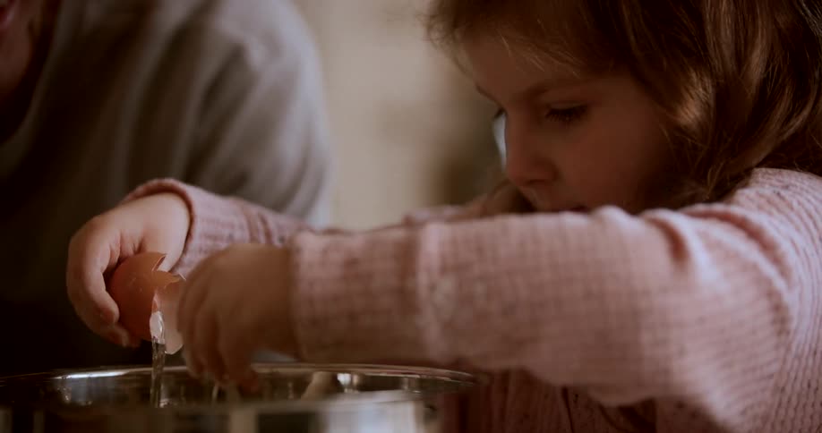 A young child wearing a pink sweater cracks an egg into a metal bowl with an adult partially visible in the background