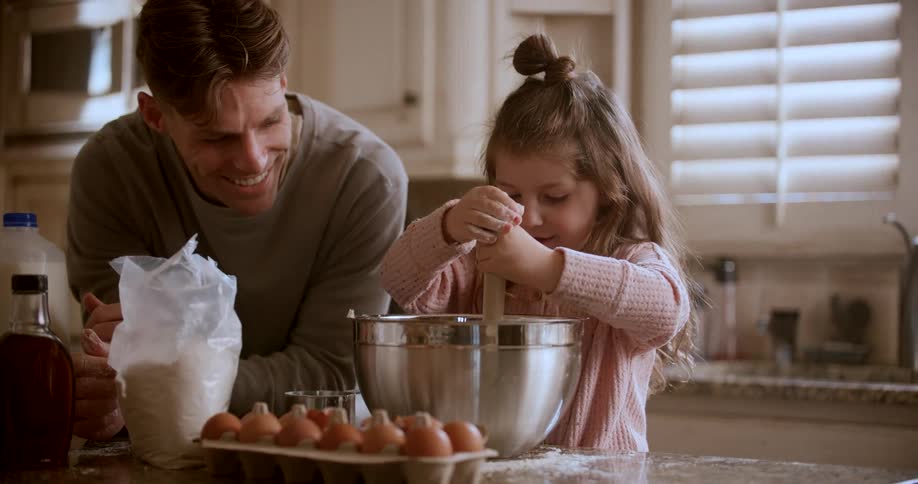 A young girl is making pancake batter in the kitchen with her father