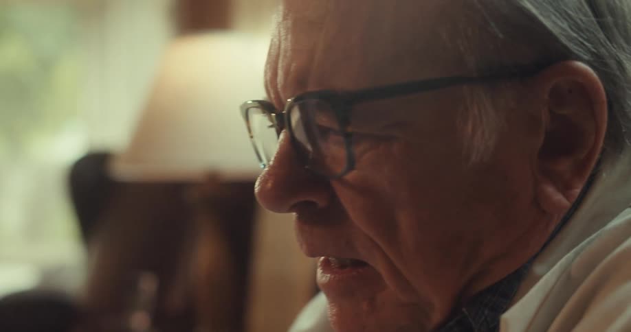 An elderly man wearing glasses and a suit sits and engages in a discussion in an office