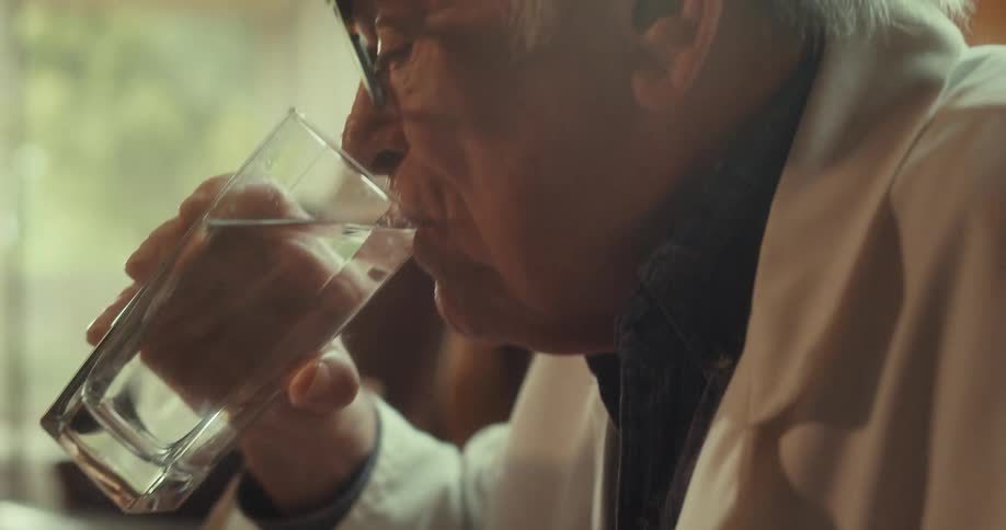 Elderly doctor drinks water from a glass cup while sitting in a living room