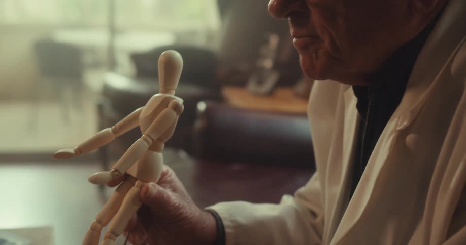 A man wearing glasses sits and talks to a client on a video call holding a doll and using it for demonstration
