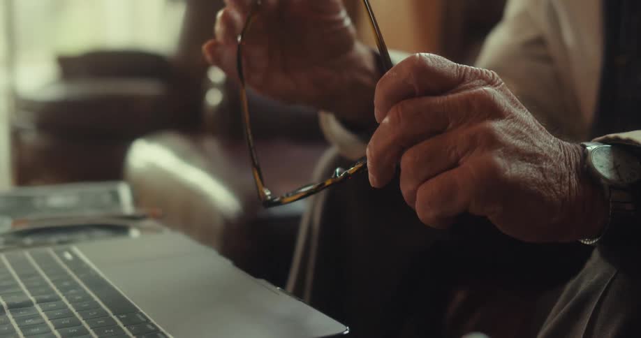 An elderly man sits in front of a computer at a table holding his eyeglasses and using hand gestures as he talks