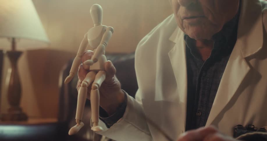 An elderly doctor holds a dummy and explains something to a patient during a video call on a computer