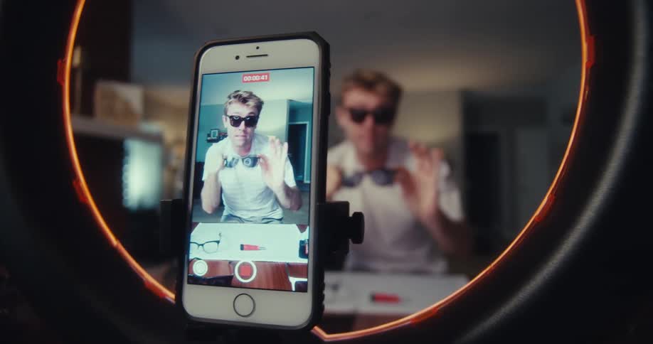 A man sits behind a ring light in his living room holding and talking about sunglasses while filming a video ad