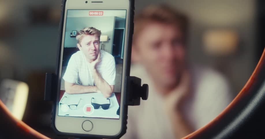 The man is talking and recording himself with his cell phone