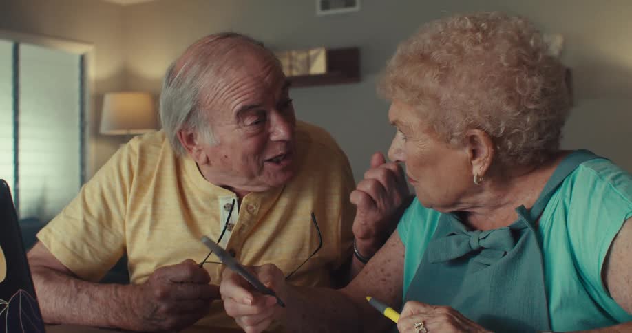 An elderly couple are talking while sitting looking at a laptop screen and holding pens in their hands