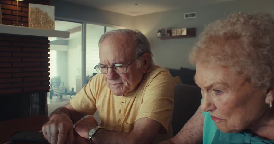 An elderly couple sitting side by side in their living room at midday scrolling through a computer on a desk with the man wearing glasses