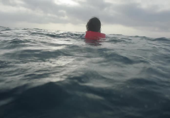 Child wearing a life vest swims through the ocean towards a group of floats under an overcast sky