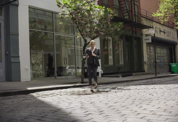 A woman in black heels holds a mobile phone while crossing to the other side of a city street at daytime