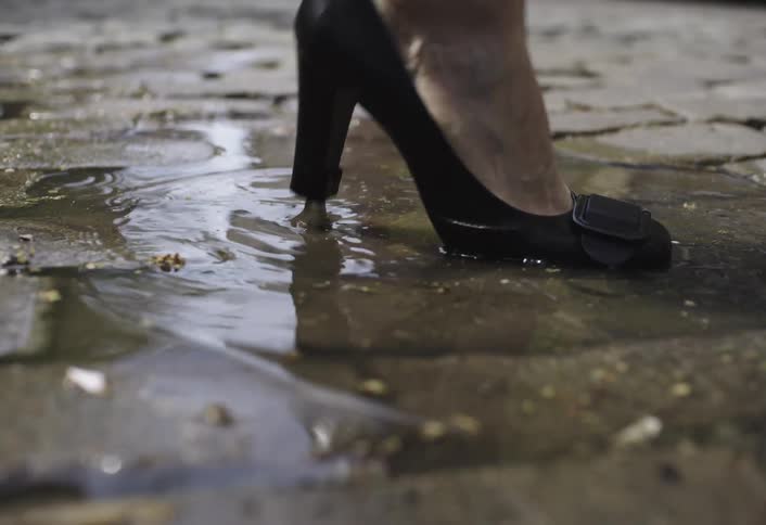 Person in black high heels walking on a city street and stepping in a puddle of water