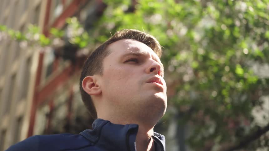 A man stands in the middle of the street beside a building under the sun with a tree nearby