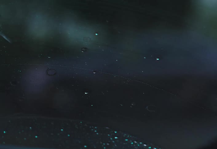 A windshield wiper clears rain from a car windshield as raindrops fall on it at night