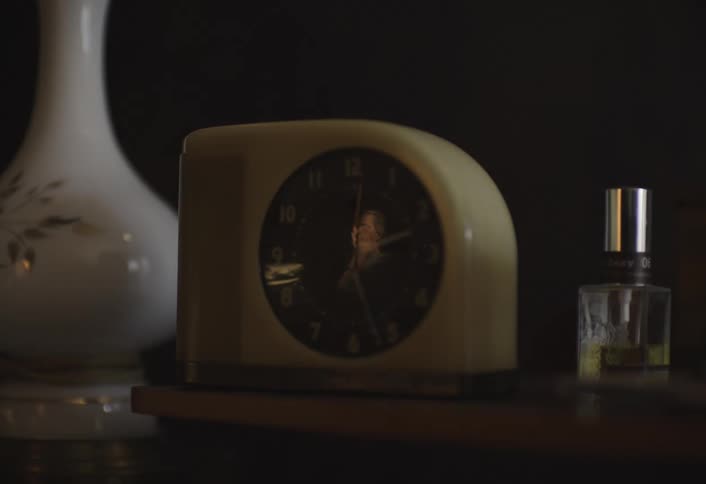 Close up of a table clock placed on a wooden surface with reflections of a woman tired and yawning visible on its glass surface