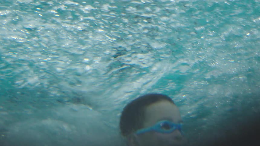 A child swims underwater wearing blue goggles surrounded by rising bubbles in a clear pool