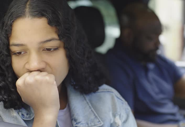 A young woman looks sad while a man in the background appears visibly angry inside a car