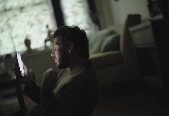 A man sits on the floor in a dimly lit room looking at his phone screen