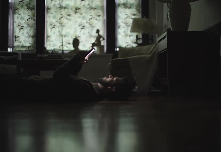 Young man sits up from lying down on the floor while texting with a phone in a dark room