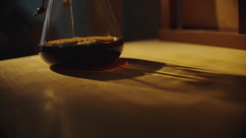 Close up of brown liquid dripping into a glass beaker placed on a wooden surface under lighting