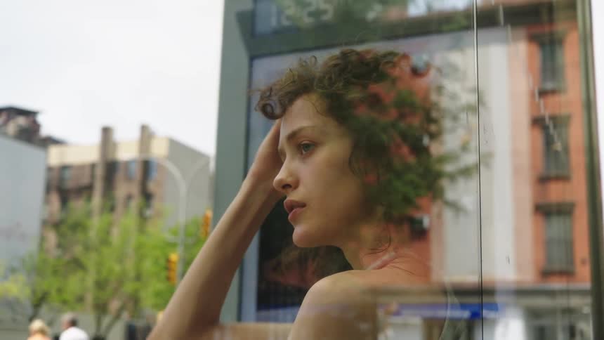 A young woman with curly hair gazes thoughtfully through a glass panel at a city street
